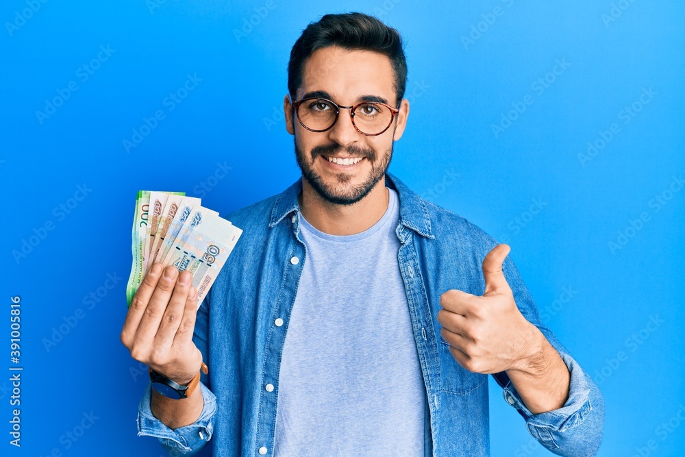 Young hispanic man holding russian ruble banknotes smiling happy and positive, thumb up doing excellent and approval sign Young hispanic man holding russian ruble banknotes smiling happy and positive, thumb up doing excellent and approval sign