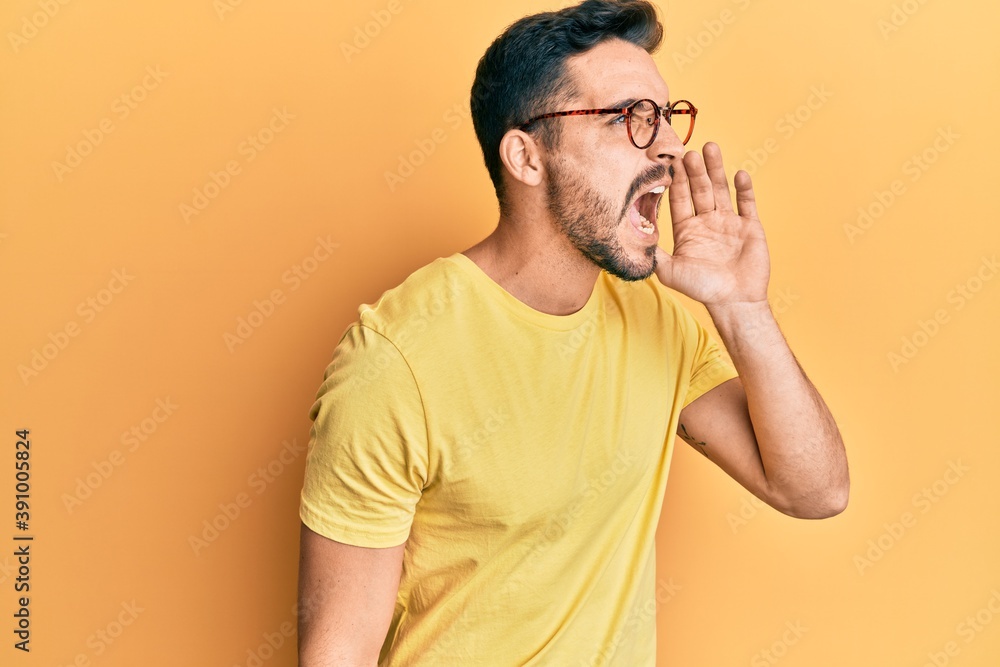Young hispanic man wearing casual clothes and glasses shouting and screaming loud to side with hand on mouth. communication concept. Young hispanic man wearing casual clothes and glasses shouting and screaming loud to side with hand on mouth. communication concept.