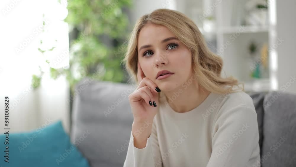 Thoughtful young woman with blond hair sitting on cozy sofa and looking around. Concept of loneliness, dreams and domestic atmosphere. Thoughtful young woman with blond hair sitting on cozy sofa and looking around. Concept of loneliness, dreams and domestic atmosphere.