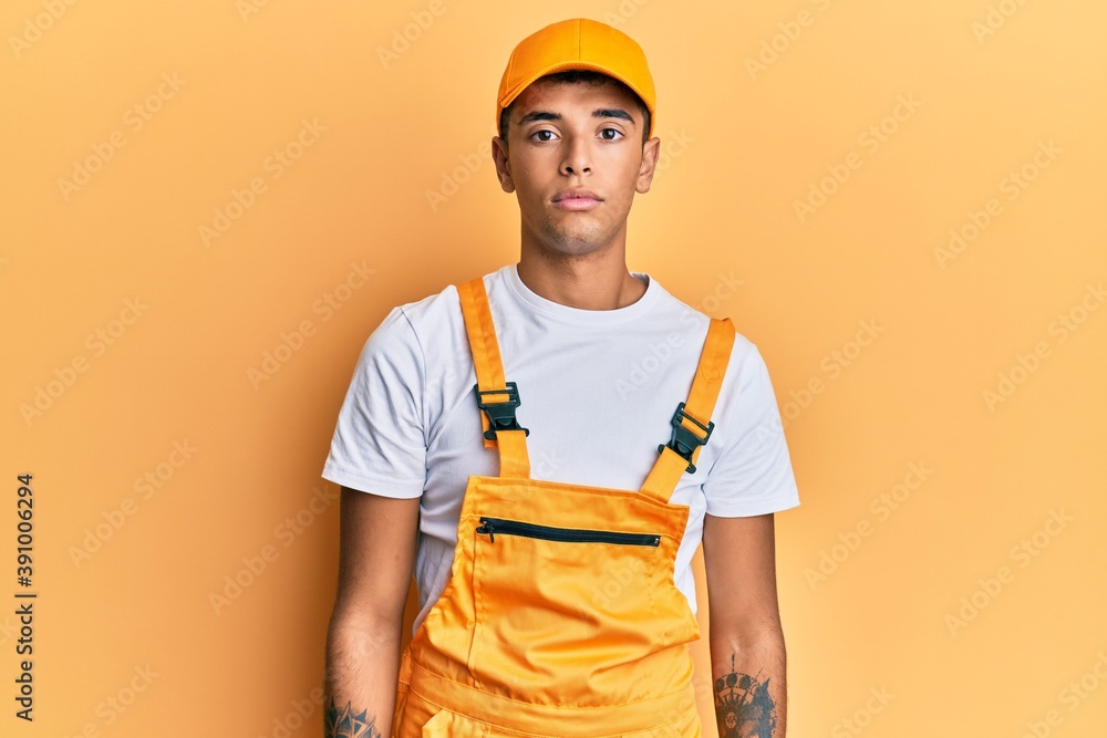 Young handsome african american man wearing handyman uniform over yellow background relaxed with serious expression on face. simple and natural looking at the camera. Young handsome african american man wearing handyman uniform over yellow background relaxed with serious expression on face. simple and natural looking at the camera.