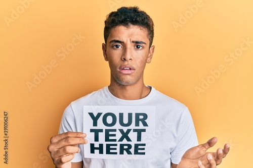 Young handsome african american man holding your text here banner clueless and confused expression. doubt concept. © Krakenimages.com