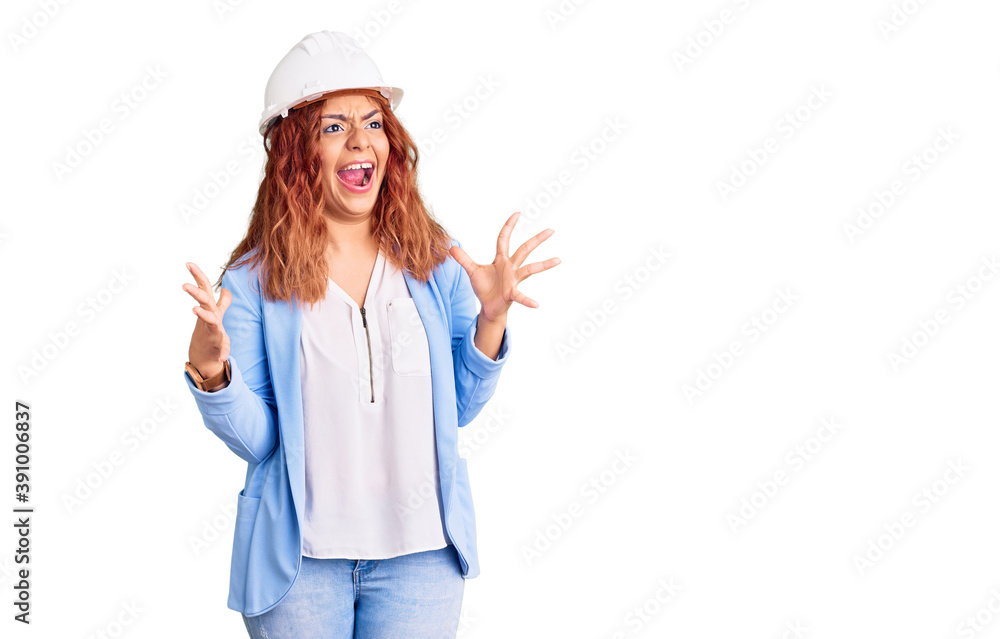 Young latin woman wearing architect hardhat crazy and mad shouting and yelling with aggressive expression and arms raised. frustration concept. Young latin woman wearing architect hardhat crazy and mad shouting and yelling with aggressive expression and arms raised. frustration concept.