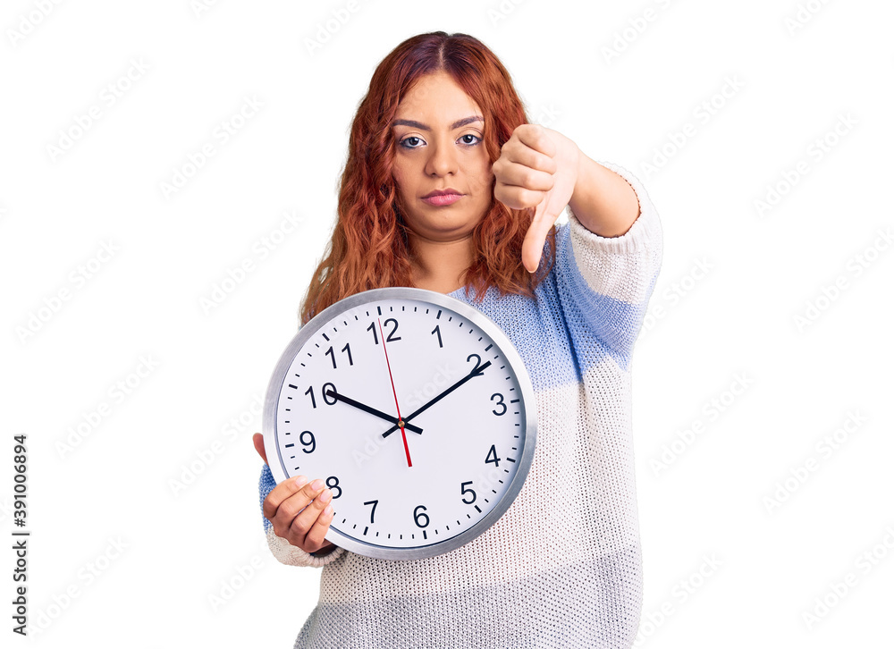 Young latin woman holding big clock with angry face, negative sign showing dislike with thumbs down, rejection concept Young latin woman holding big clock with angry face, negative sign showing dislike with thumbs down, rejection concept