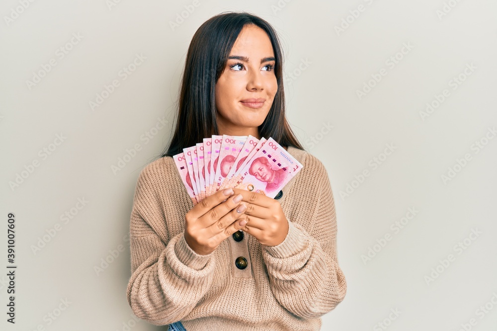 Young beautiful hispanic girl holding yuan chinese banknotes smiling looking to the side and staring away thinking. Young beautiful hispanic girl holding yuan chinese banknotes smiling looking to the side and staring away thinking.