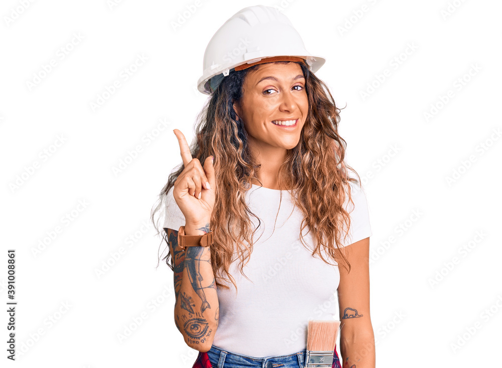 Young hispanic woman with tattoo wearing hardhat and builder clothes showing and pointing up with finger number one while smiling confident and happy. Young hispanic woman with tattoo wearing hardhat and builder clothes showing and pointing up with finger number one while smiling confident and happy.