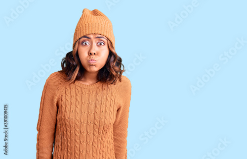 Young beautiful mixed race woman wearing wool sweater and winter hat puffing cheeks with funny face. mouth inflated with air, crazy expression. photo