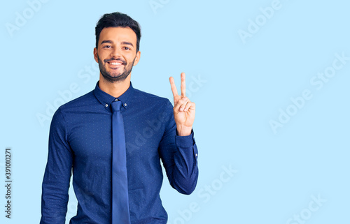 Young handsome hispanic man wearing business clothes smiling with happy face winking at the camera doing victory sign. number two.