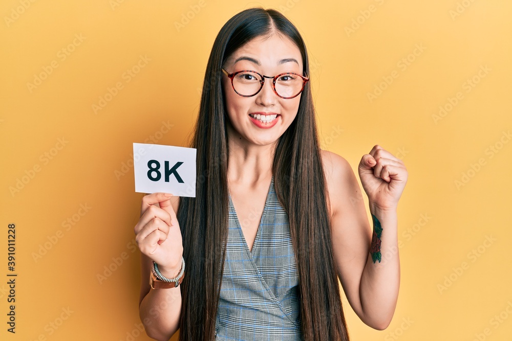 Young chinese woman holding 8k resolution technology paper screaming proud, celebrating victory and success very excited with raised arm Young chinese woman holding 8k resolution technology paper screaming proud, celebrating victory and success very excited with raised arm
