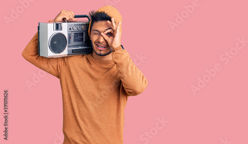 Handsome latin american young man holding boombox, listening to music doing ok gesture with hand smiling, eye looking through fingers with happy face.