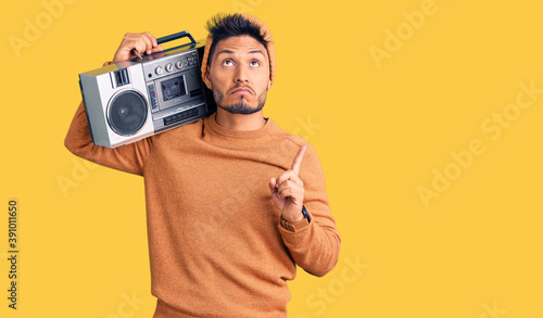 Handsome latin american young man holding boombox, listening to music pointing up looking sad and upset, indicating direction with fingers, unhappy and depressed.