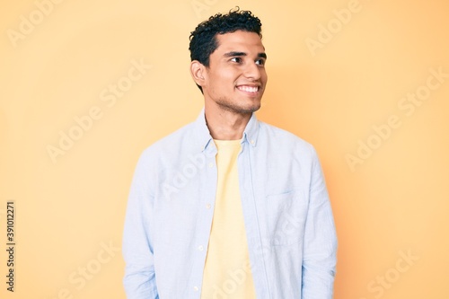 Young handsome hispanic man standing over yellow background looking to side, relax profile pose with natural face and confident smile.