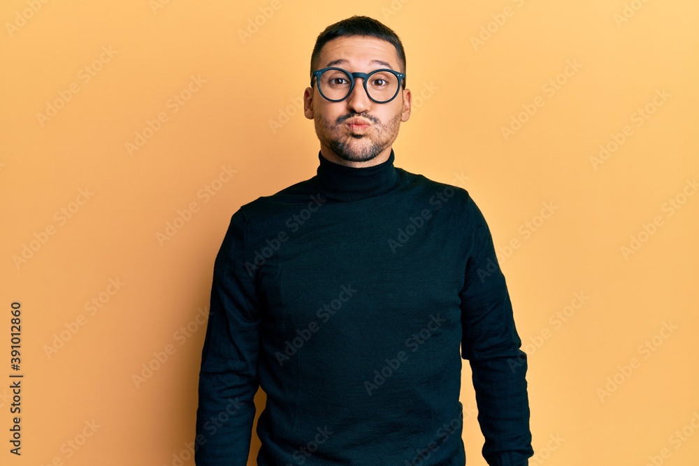 Handsome man with tattoos wearing turtleneck sweater and glasses puffing cheeks with funny face. mouth inflated with air, crazy expression. Handsome man with tattoos wearing turtleneck sweater and glasses puffing cheeks with funny face. mouth inflated with air, crazy expression.