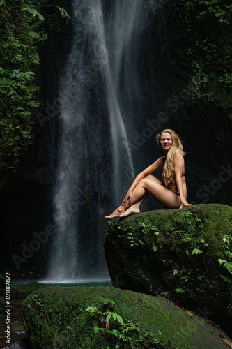 Young Caucasian woman with long blond hair sitting on the rock and enjoiyng waterfall landscape. Travel lifestyle. Leke Leke waterfall, Bali.