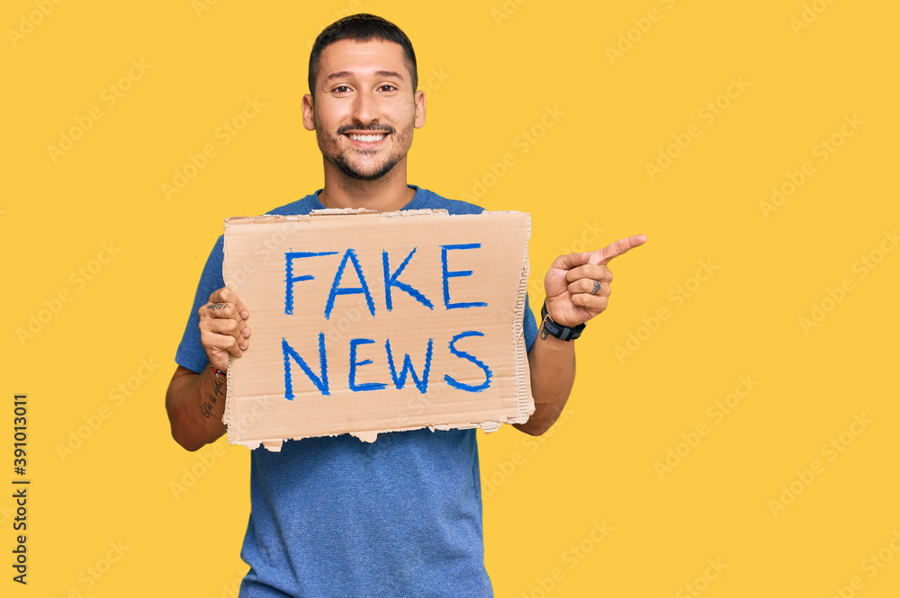 Handsome man with tattoos holding fake news banner smiling happy pointing with hand and finger to the side Handsome man with tattoos holding fake news banner smiling happy pointing with hand and finger to the side