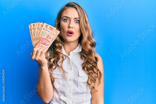 Young blonde girl holding mexican pesos scared and amazed with open mouth for surprise, disbelief face photo