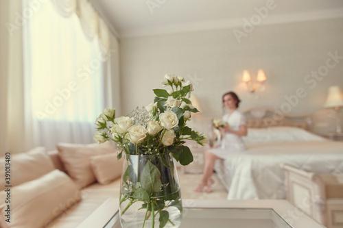 A girl in a white negligee is sitting on the bed with a bouquet of white roses in her hand