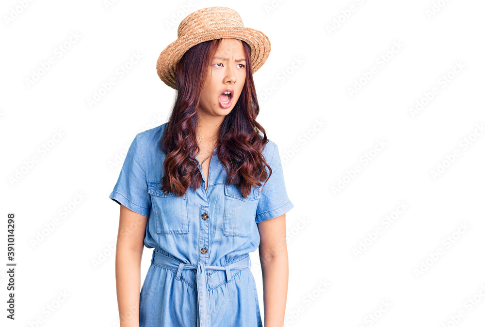 Young beautiful chinese girl wearing summer hat angry and mad screaming frustrated and furious, shouting with anger. rage and aggressive concept.