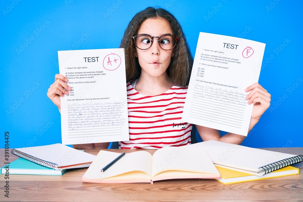 Cute hispanic girl showing failed and passe exam sitting on the table making fish face with mouth and squinting eyes, crazy and comical. Cute hispanic girl showing failed and passe exam sitting on the table making fish face with mouth and squinting eyes, crazy and comical.