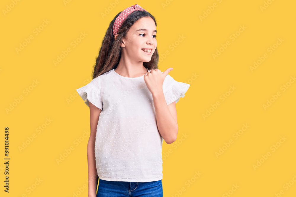 Cute hispanic child girl wearing casual white tshirt smiling with happy face looking and pointing to the side with thumb up. Cute hispanic child girl wearing casual white tshirt smiling with happy face looking and pointing to the side with thumb up.