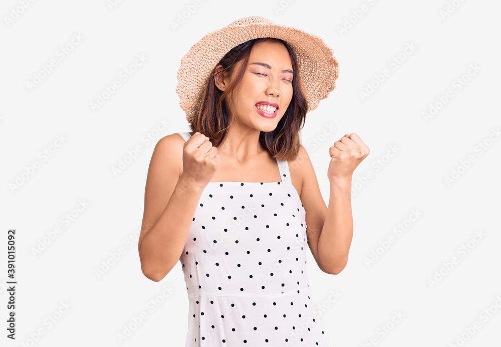 Young beautiful chinese girl wearing summer hat excited for success with arms raised and eyes closed celebrating victory smiling. winner concept. Young beautiful chinese girl wearing summer hat excited for success with arms raised and eyes closed celebrating victory smiling. winner concept.