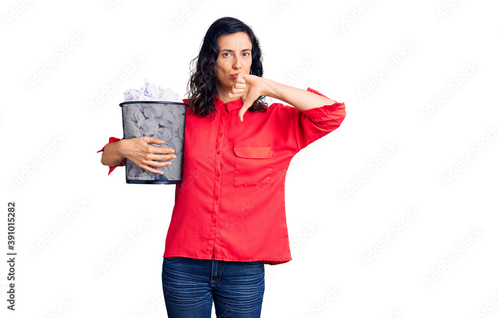 Young beautiful hispanic woman holding paper bin full of crumpled papers with angry face, negative sign showing dislike with thumbs down, rejection concept Young beautiful hispanic woman holding paper bin full of crumpled papers with angry face, negative sign showing dislike with thumbs down, rejection concept