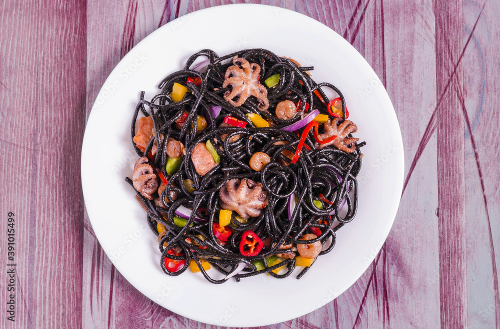 black spaghetti with seafood and vegetables on a white plate decorated with red pepper wooden background top view