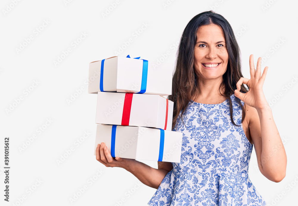 Young beautiful brunette woman holding birthday gift doing ok sign with fingers, smiling friendly gesturing excellent symbol Young beautiful brunette woman holding birthday gift doing ok sign with fingers, smiling friendly gesturing excellent symbol