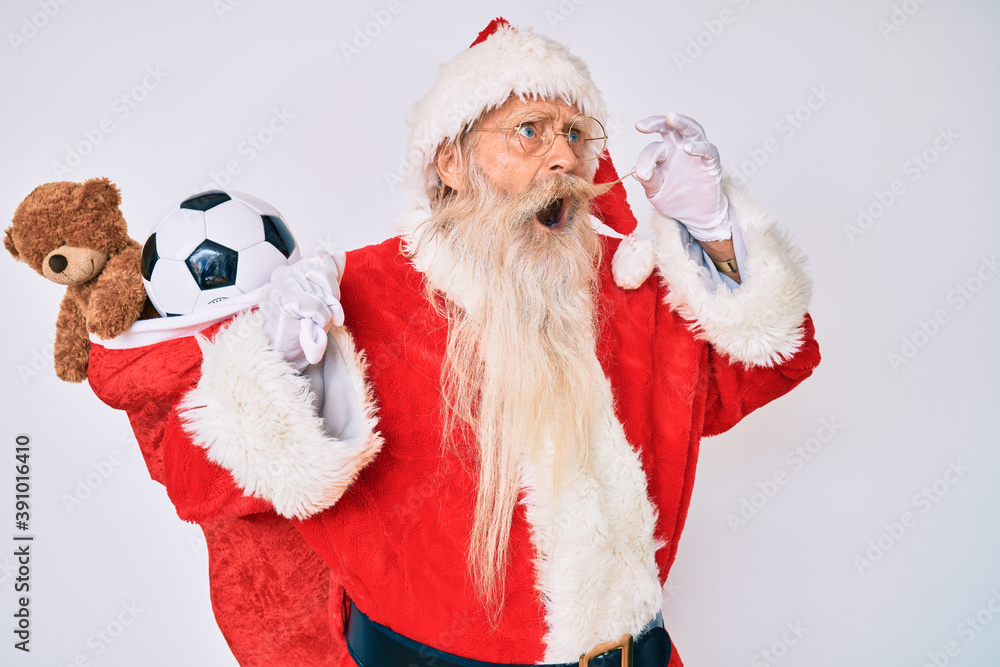 Old senior man with grey hair and long beard wearing santa claus costume holding mustache angry and mad screaming frustrated and furious, shouting with anger. rage and aggressive concept. Old senior man with grey hair and long beard wearing santa claus costume holding mustache angry and mad screaming frustrated and furious, shouting with anger. rage and aggressive concept.