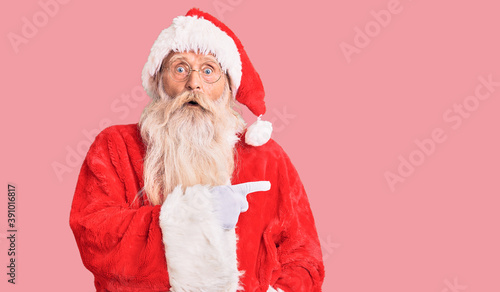 Old senior man with grey hair and long beard wearing traditional santa claus costume surprised pointing with finger to the side, open mouth amazed expression.