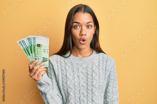 Beautiful hispanic woman holding 10000 south korean won banknotes scared and amazed with open mouth for surprise, disbelief face