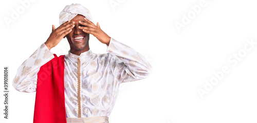 African handsome man wearing tradition sherwani saree clothes covering eyes with hands smiling cheerful and funny. blind concept.
