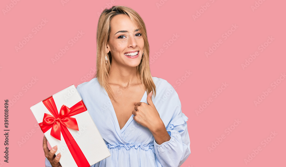 Beautiful blonde woman holding gift pointing finger to one self smiling happy and proud Beautiful blonde woman holding gift pointing finger to one self smiling happy and proud