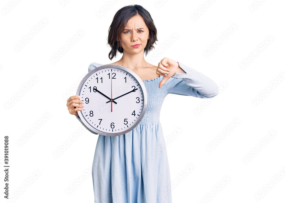 Young beautiful girl holding big clock with angry face, negative sign showing dislike with thumbs down, rejection concept Young beautiful girl holding big clock with angry face, negative sign showing dislike with thumbs down, rejection concept