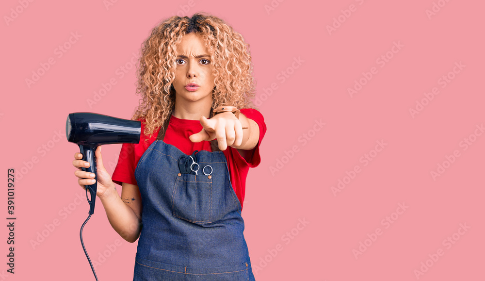Young blonde woman with curly hair wearing hairdresser apron and holding dryer blow pointing with finger to the camera and to you, confident gesture looking serious Young blonde woman with curly hair wearing hairdresser apron and holding dryer blow pointing with finger to the camera and to you, confident gesture looking serious