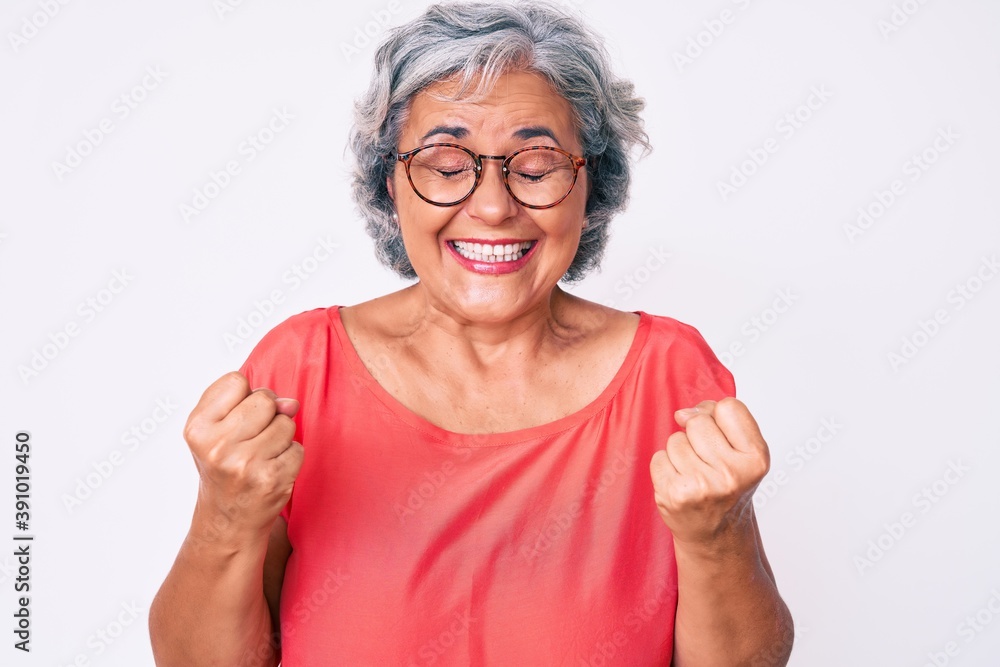 Senior hispanic grey- haired woman wearing casual clothes and glasses very happy and excited doing winner gesture with arms raised, smiling and screaming for success. celebration concept. Senior hispanic grey- haired woman wearing casual clothes and glasses very happy and excited doing winner gesture with arms raised, smiling and screaming for success. celebration concept.