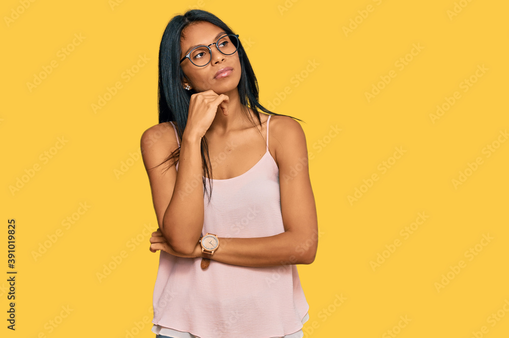 Young african american woman wearing casual clothes and glasses with hand on chin thinking about question, pensive expression. smiling with thoughtful face. doubt concept. Young african american woman wearing casual clothes and glasses with hand on chin thinking about question, pensive expression. smiling with thoughtful face. doubt concept.