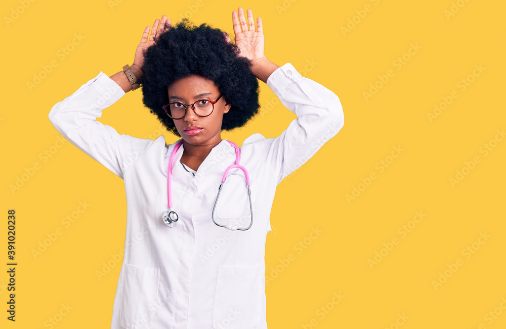 Young african american woman wearing doctor coat and stethoscope doing bunny ears gesture with hands palms looking cynical and skeptical. easter rabbit concept.