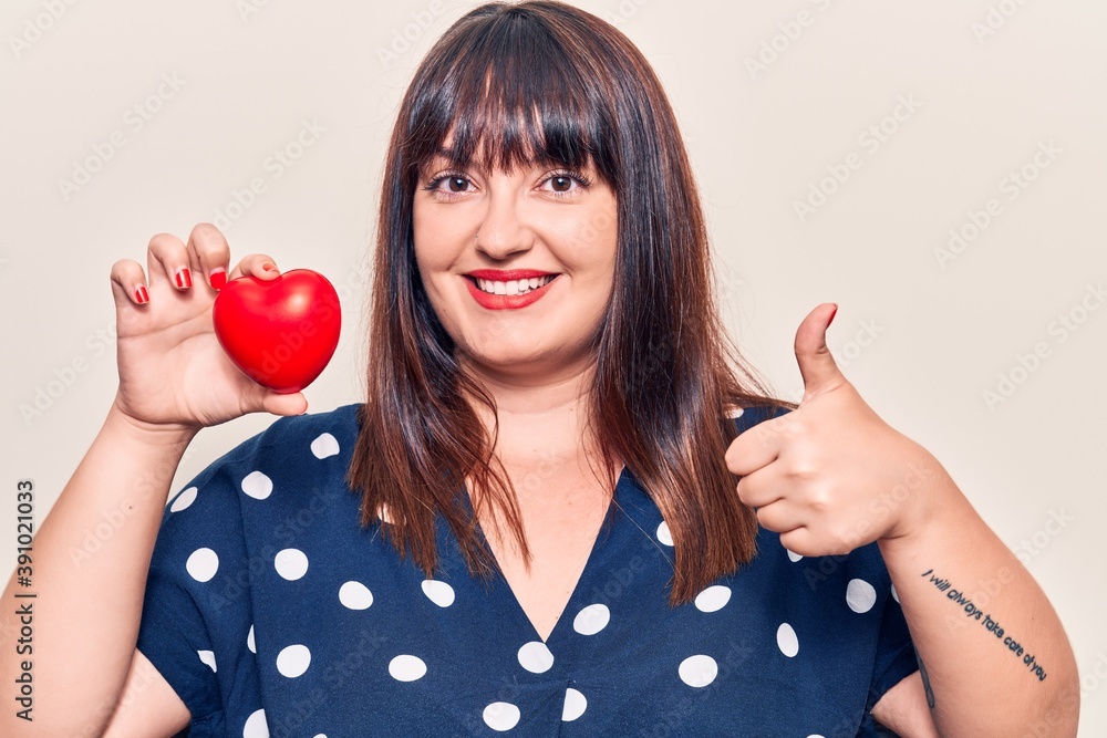 Young plus size woman holding heart smiling happy and positive, thumb up doing excellent and approval sign Young plus size woman holding heart smiling happy and positive, thumb up doing excellent and approval sign