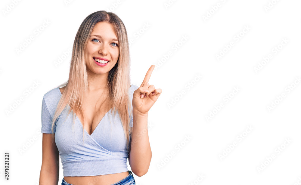 Young beautiful blonde woman wearing casual clothes showing and pointing up with finger number one while smiling confident and happy. Young beautiful blonde woman wearing casual clothes showing and pointing up with finger number one while smiling confident and happy.