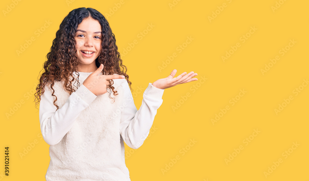Beautiful kid girl with curly hair wearing casual clothes amazed and smiling to the camera while presenting with hand and pointing with finger. Beautiful kid girl with curly hair wearing casual clothes amazed and smiling to the camera while presenting with hand and pointing with finger.