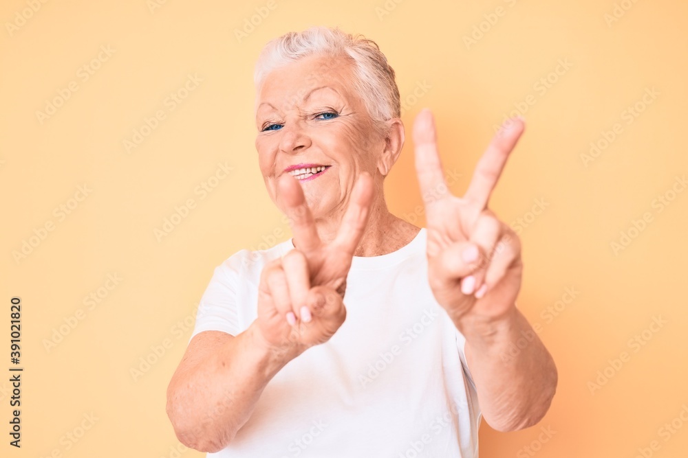 Senior beautiful woman with blue eyes and grey hair wearing classic white tshirt over yellow background smiling looking to the camera showing fingers doing victory sign. number two. Senior beautiful woman with blue eyes and grey hair wearing classic white tshirt over yellow background smiling looking to the camera showing fingers doing victory sign. number two.