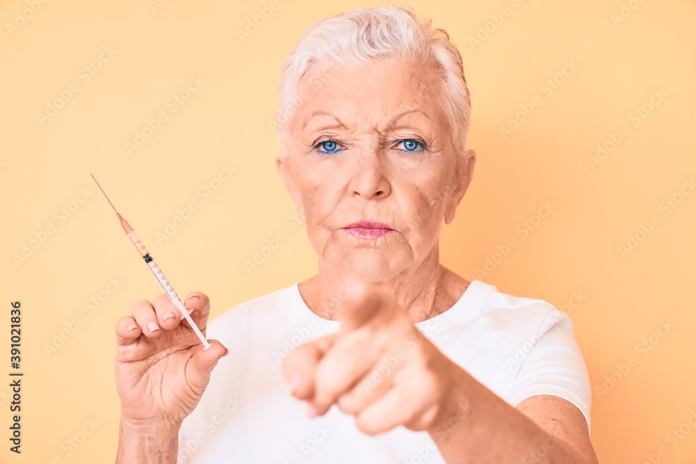 Senior beautiful woman with blue eyes and grey hair holding syringe pointing with finger to the camera and to you, confident gesture looking serious Senior beautiful woman with blue eyes and grey hair holding syringe pointing with finger to the camera and to you, confident gesture looking serious