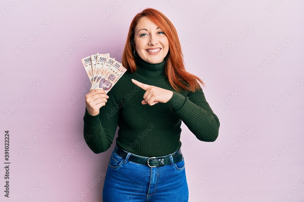 Beautiful redhead woman holding 500 mexican pesos banknotes smiling happy pointing with hand and finger Beautiful redhead woman holding 500 mexican pesos banknotes smiling happy pointing with hand and finger