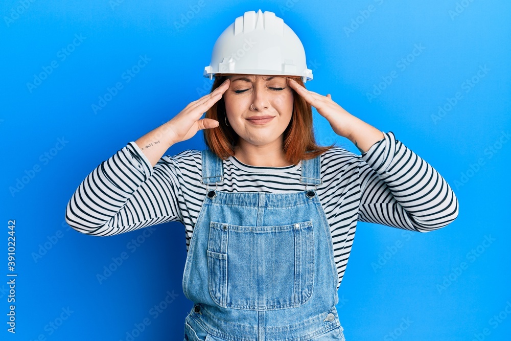 Beautiful redhead woman wearing architect hardhat with hand on head, headache because stress. suffering migraine.