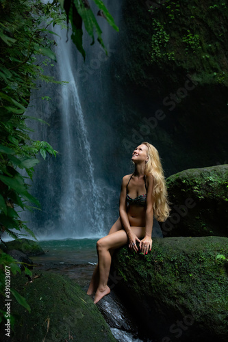 Young Caucasian woman with long blond hair sitting on the rock and enjoiyng waterfall landscape. Travel lifestyle. Leke Leke waterfall  Bali.