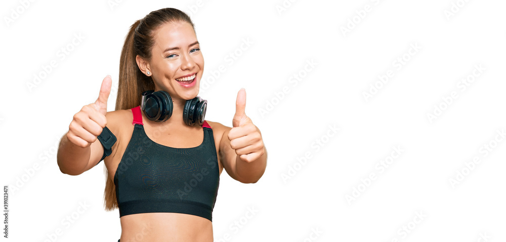 Beautiful blonde woman wearing gym clothes and using headphones approving doing positive gesture with hand, thumbs up smiling and happy for success. winner gesture. Beautiful blonde woman wearing gym clothes and using headphones approving doing positive gesture with hand, thumbs up smiling and happy for success. winner gesture.