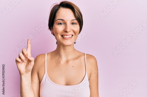 Young brunette woman with short hair over pink background pointing finger up with successful idea. exited and happy. number one.