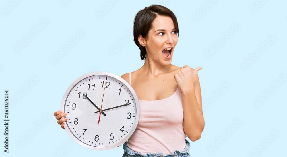 Young brunette woman with short hair holding big clock pointing thumb up to the side smiling happy with open mouth Young brunette woman with short hair holding big clock pointing thumb up to the side smiling happy with open mouth