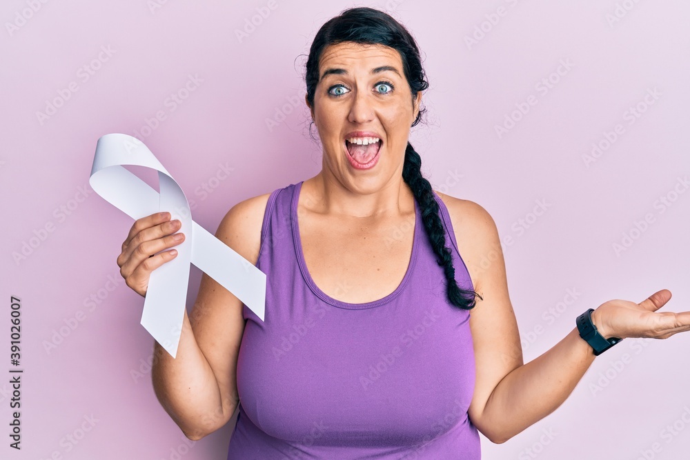 Plus size brunette woman holding white ribbon celebrating achievement with happy smile and winner expression with raised hand Plus size brunette woman holding white ribbon celebrating achievement with happy smile and winner expression with raised hand