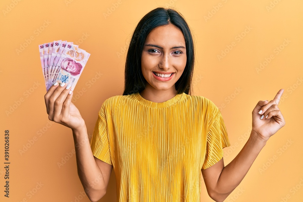 Young brunette woman holding 50 mexican pesos banknotes smiling happy pointing with hand and finger to the side Young brunette woman holding 50 mexican pesos banknotes smiling happy pointing with hand and finger to the side